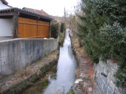 Bach mit hart verbauten Ufern (verfugte Steine); Betonmauer und Gebäude direkt am Ufer im linken Bildbereiche; Steinmauer und hohe Fichten direkt am Ufer. Bach mit hart verbauten Ufern (verfugte Steine); Betonmauer und Gebäude direkt am Ufer im linken Bildbereiche; Steinmauer und hohe Fichten direkt am Ufer.
